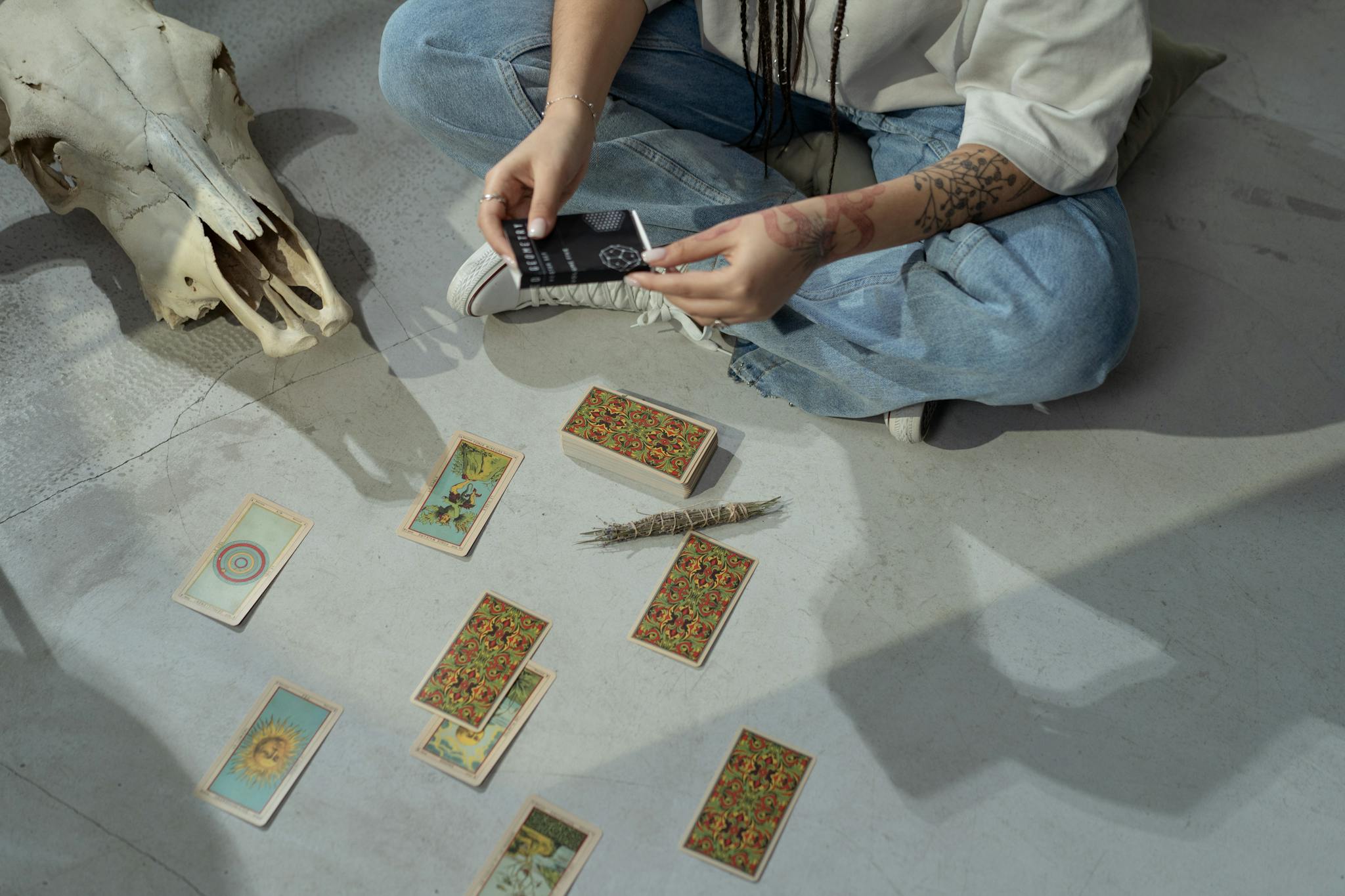 A tattooed person sits on the floor performing a tarot reading with cards and a skull.